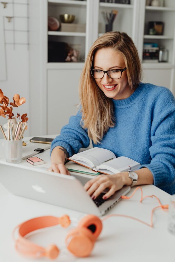 pexels-photo-5908728 A cheerful woman in a blue sweater working remotely with a laptop in a cozy home setting.